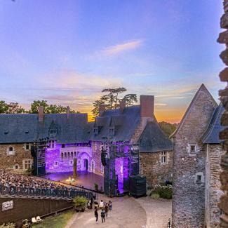 Vue large sur la cour du château du Plessis-Macé, avec la scène de plein air installée entre les logis historiques et le gradin rempli de spectateurs. Dans une atmosphère de fin de journée, un ciel aux couleurs coucher de soleil.