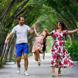 Un homme et une femme avancent sous une allée végétale, rigolent et tiennent par la main leur petite fille, en train de sauter entre eux deux.