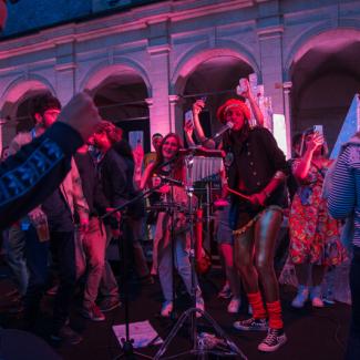Groupe de chanteurs et musiciens. Photo de nuit dans un cloître, éclairages avec des spots de couleurs rouges et roses. Des personnes qui dansent autour des musiciens.