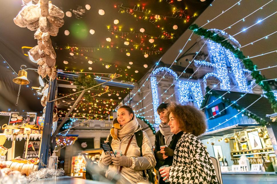 Trois femmes devant un étal sur le marché de Noël à Angers, avec des illuminations sur le chalet et le Grand théâtre en arrière-plan.